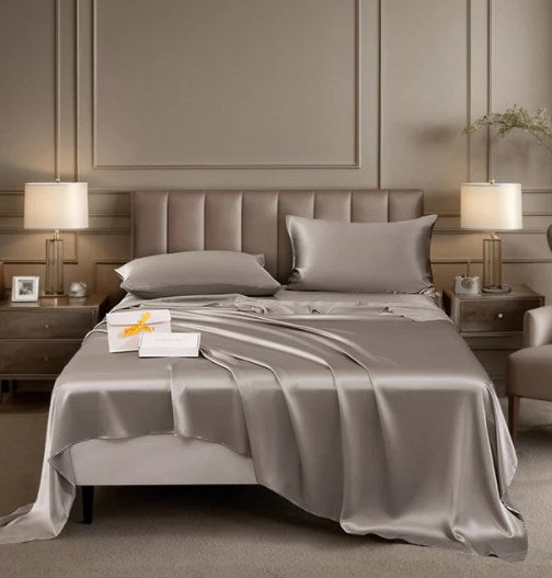 A luxurious silver-grey silk bedding set, including a flat sheet and pillowcases, elegantly draped on a bed in a sophisticated bedroom setting