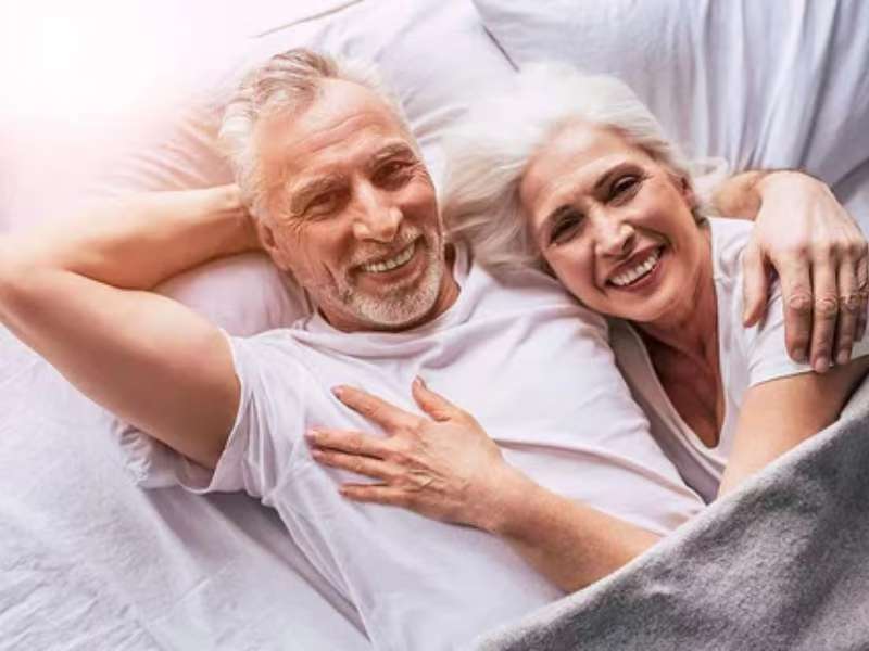 Senior couple smiling happily while lying together on white bedding, showcasing comfort and satisfaction with their sleep experience