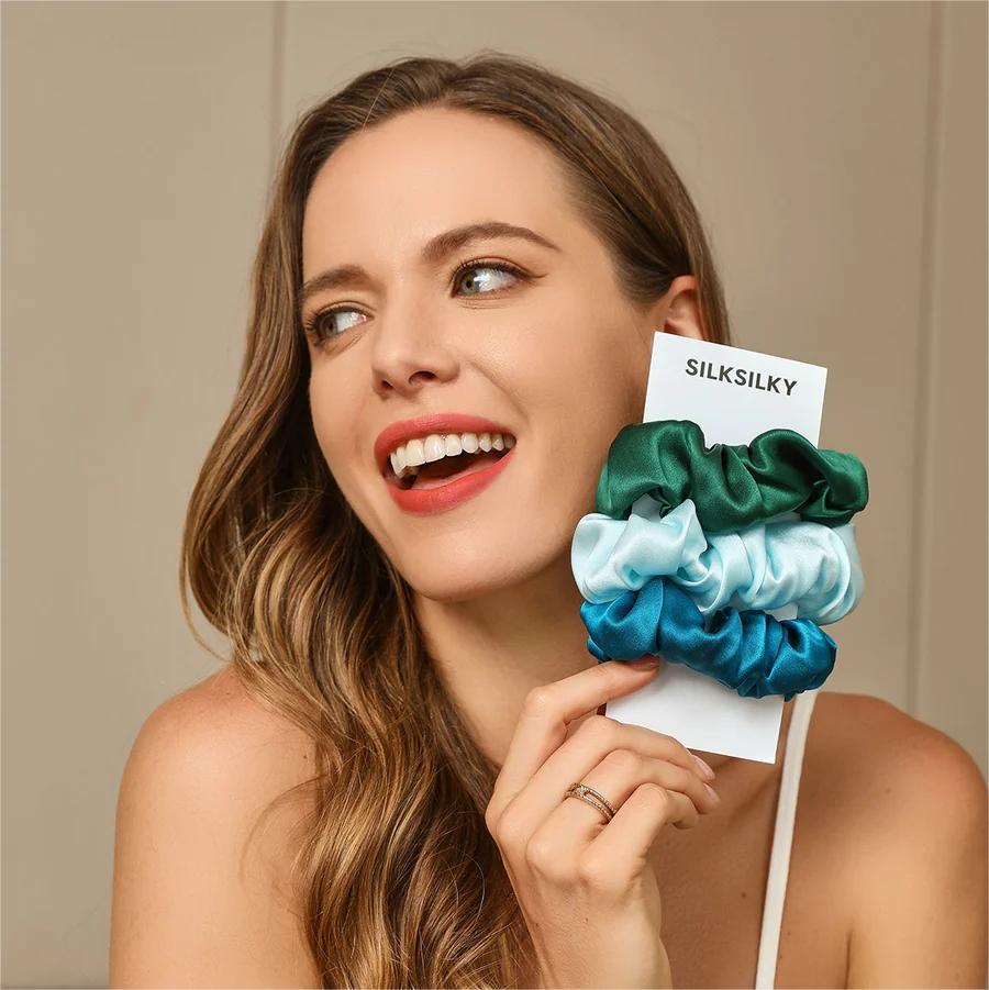 A smiling woman holding a set of three silk scrunchies in shades of green and blue