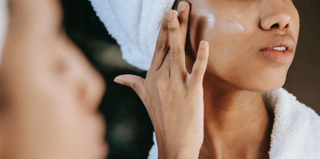 Woman applying facial skincare cream while looking in mirror wearing white towel robe
