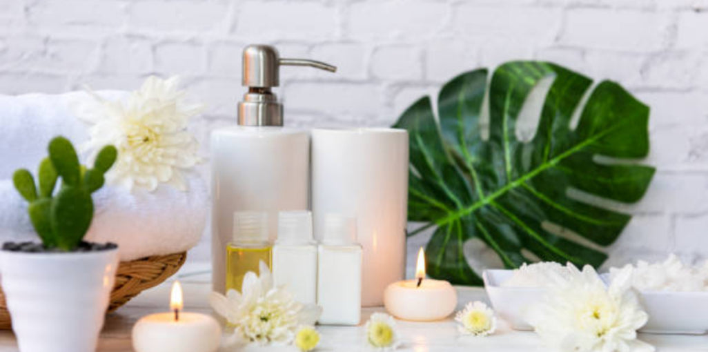 Spa bathroom setup with white ceramic soap dispenser, small potted cactus, white candles, essential oil bottles, monstera leaf, and white flowers against a white brick wall background