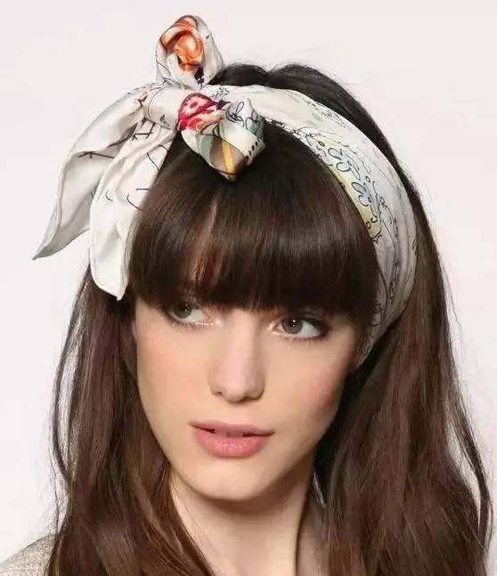 A woman with long brown hair and bangs wears a light-colored, patterned scarf as a head wrap, tied in a prominent bow-like knot on top of her head