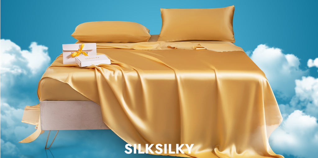 Luxurious golden silk bed sheets and pillowcases by SILKSILKY displayed on a white bed against a blue sky background with clouds. A decorative gift box with a yellow ribbon sits on the bed, highlighting the premium nature of the product